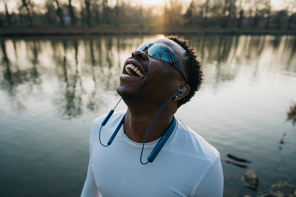 Man laughing by a lake wearing blue neckband earbuds and sunglasses.