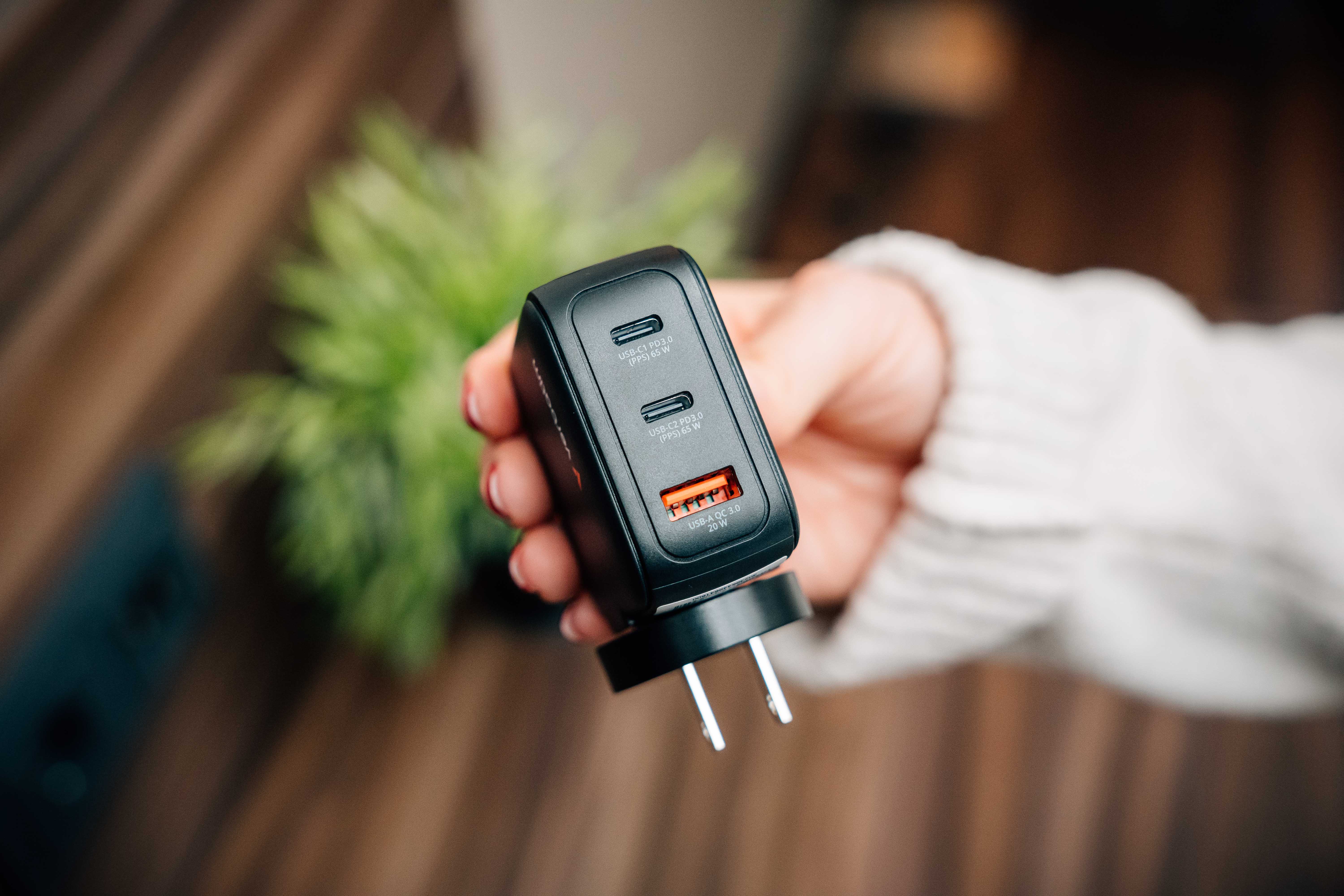 Jemand hält ein GaN-Ladegerät in der Hand, an dessen Ende die USB-C- und USB-A-Stecker zu sehen sind. Im Hintergrund ist eine Pflanze.