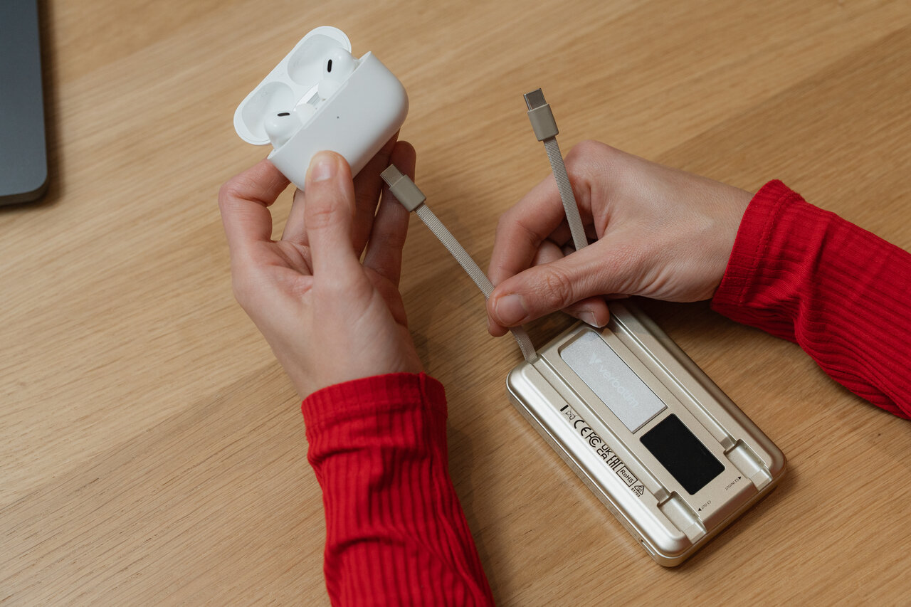 En guldfärgad powerbank på ett bord. En hand som visar de två inbyggda USB-C-kablarna, varav den ena är ansluten till ett AirPods-fodral.