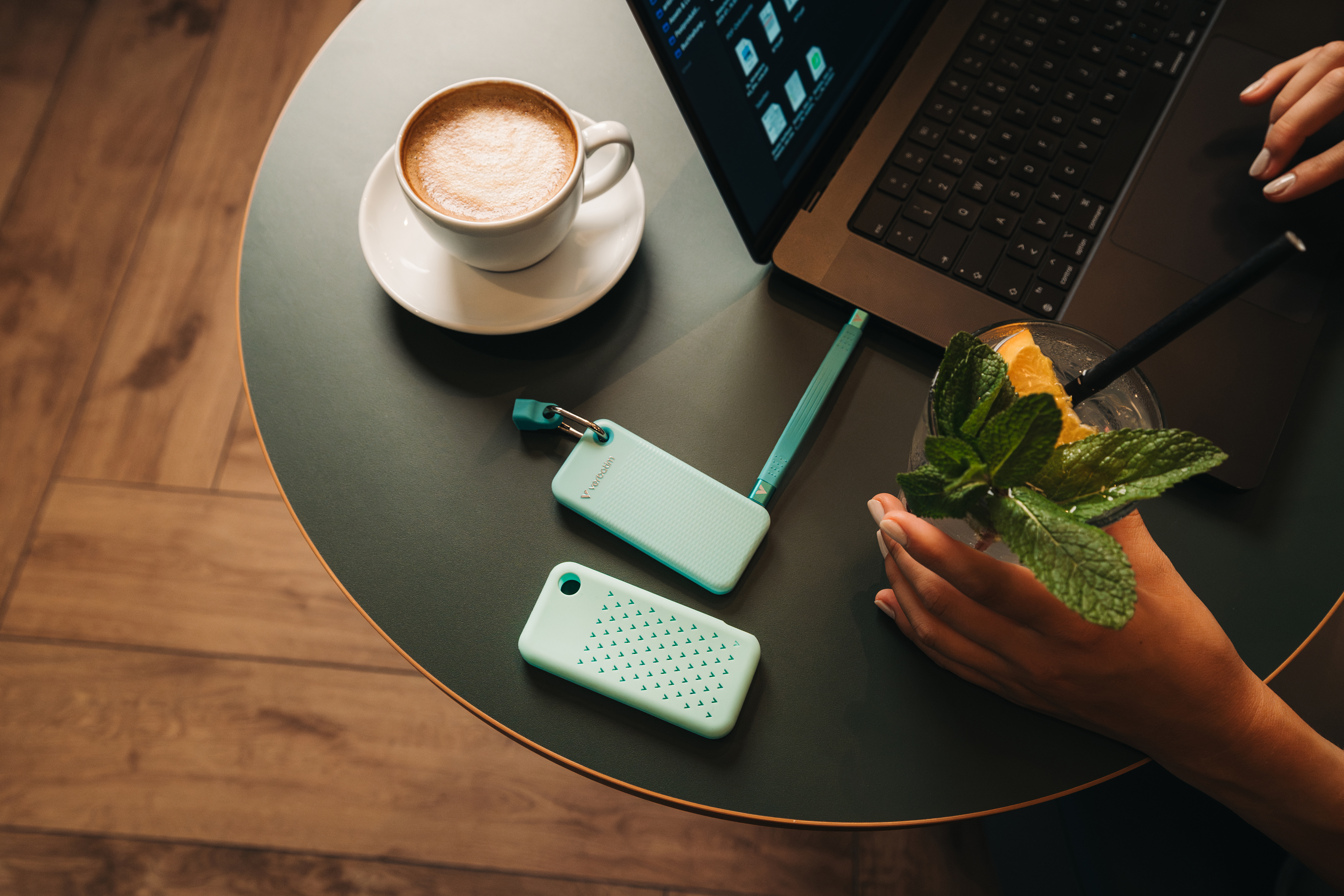 Eine arktisblaue Verbatim Pocket SSD steckt in einem Laptop auf einem Tisch in einem Café. Dazu gibt's eine Tasse Kaffee und ein Getränk mit Minzblättchen.