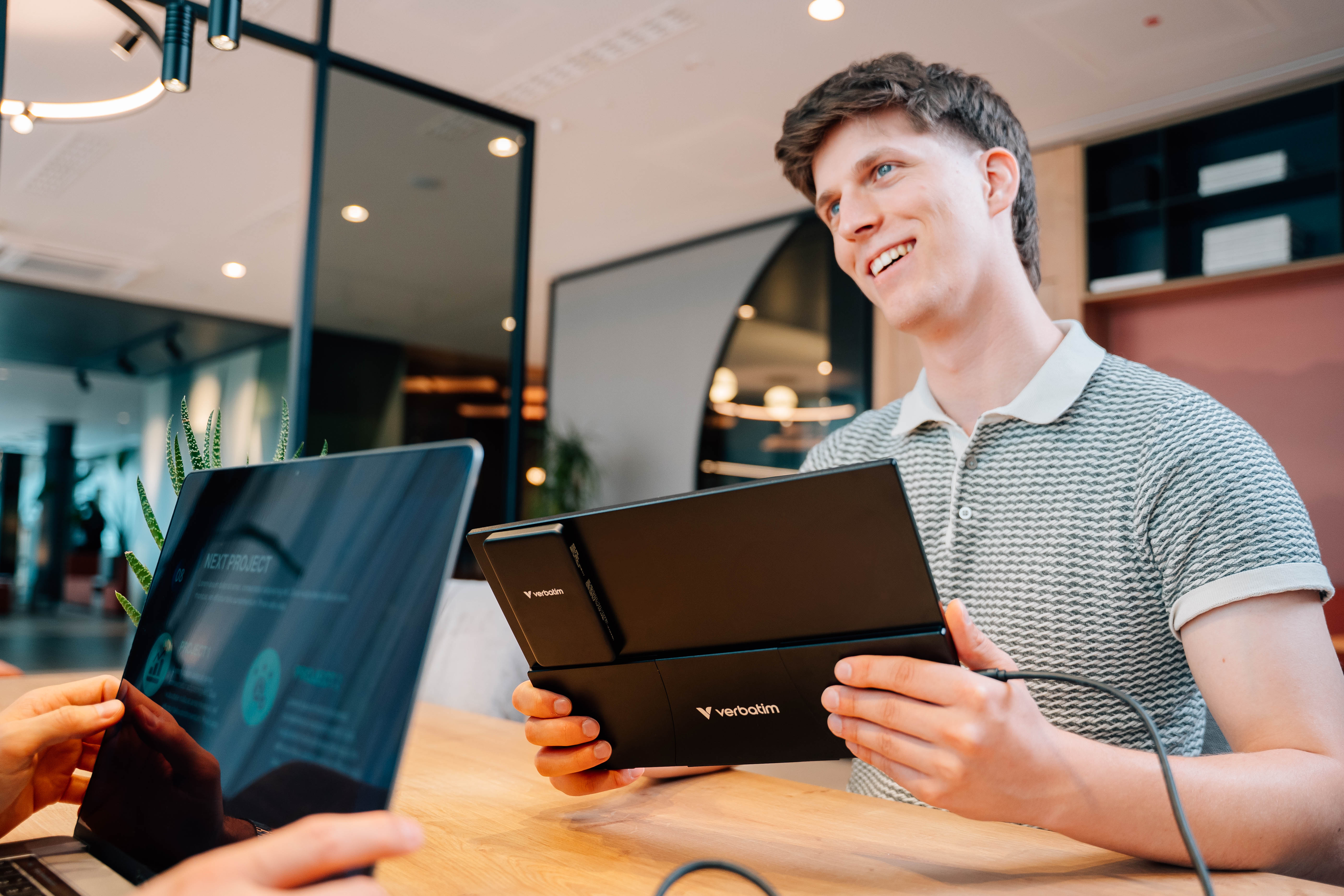 Ein Typ mit einem schwarzen tragbaren Monitor von Verbatim gegenüber einer anderen Person mit einem Laptop.